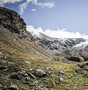 Paysage alpin sauvage avec un vallon rocheux et herbeux menant vers des sommets enneigés sous un ciel partiellement nuageux. Ambiance de haute montagne en automne, terrain minéral et végétation rase.