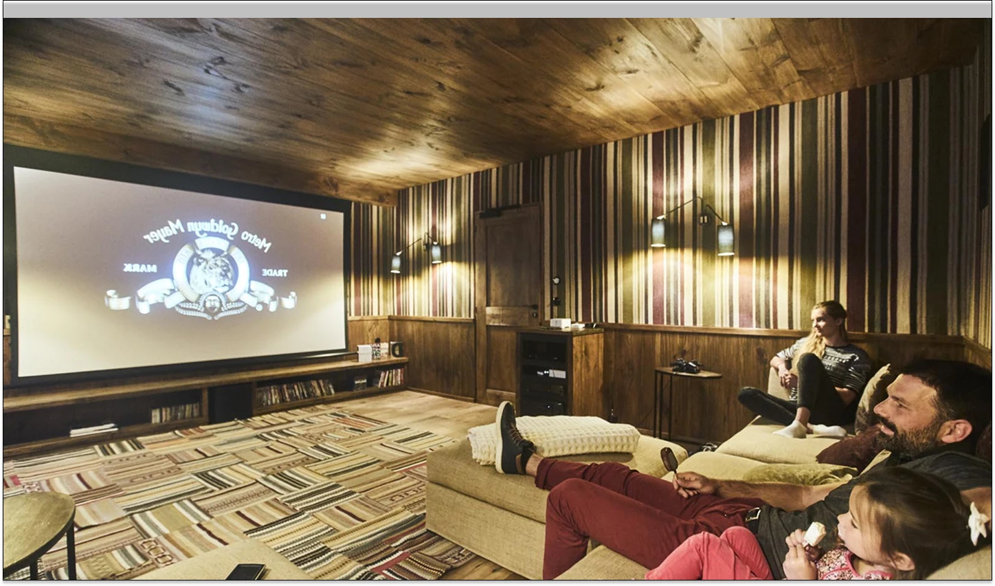 Salle de cinéma privée en chalet avec plafond et murs en bois, habillés de lames verticales multicolores. Une famille est allongée sur des canapés confortables face à un grand écran de projection. Ambiance chaleureuse et cosy avec un éclairage tamisé.