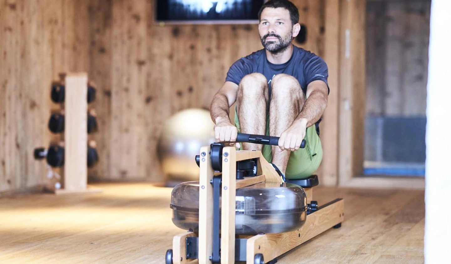 Homme s'entraînant sur un rameur en bois dans une salle de fitness en chalet alpin. Murs en bois de mélèze, haltères et ballon de fitness visibles en arrière-plan. Ambiance salle de sport haut de gamme et naturelle.