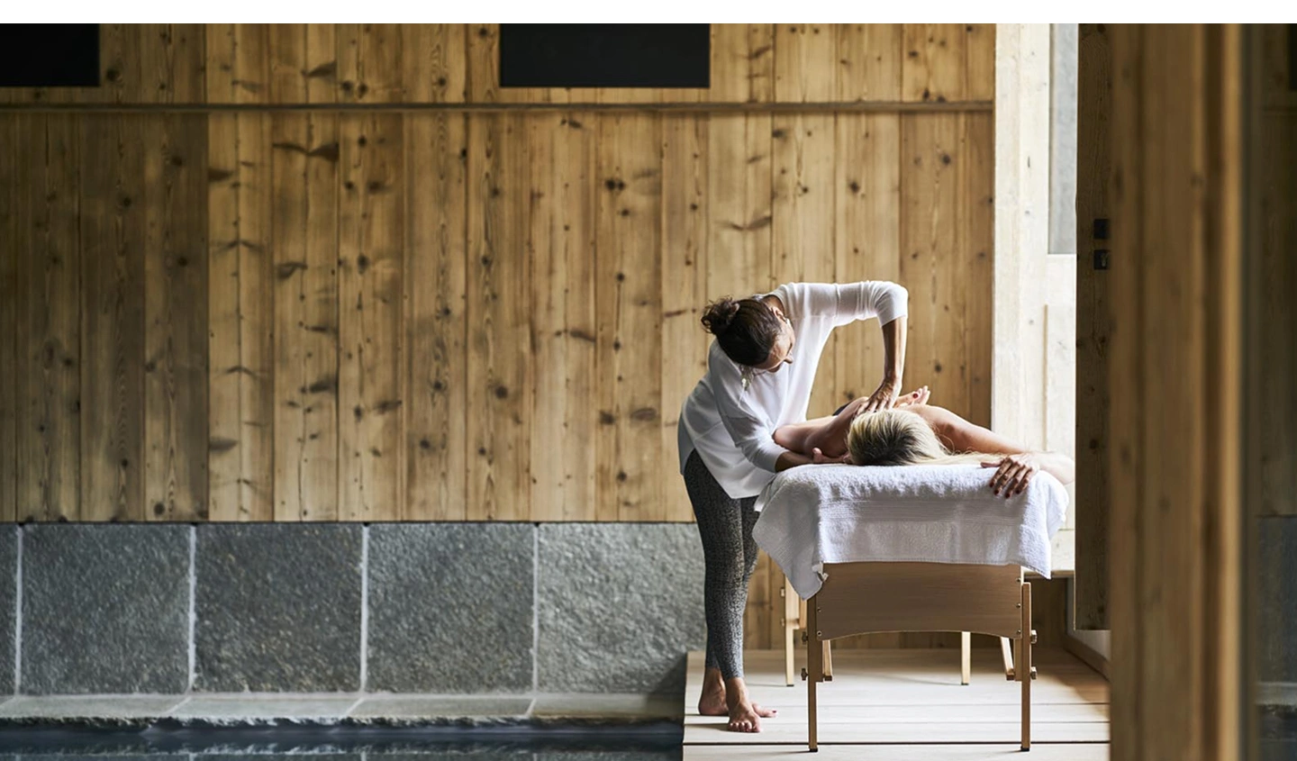 Masseuse prodiguant un soin du dos à une cliente allongée sur une table de massage drapée d'une serviette blanche. Cadre spa en chalet alpin avec mur en bois de mélèze, soubassement en pierre de taille et lumière naturelle entrant par une baie vitrée.